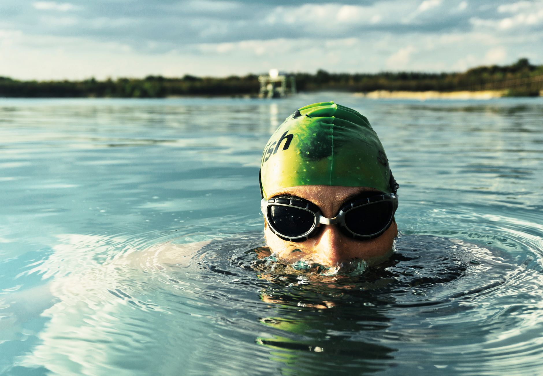 A determined swimmer in goggles and cap emerges from a tranquil lake on a sunny day.