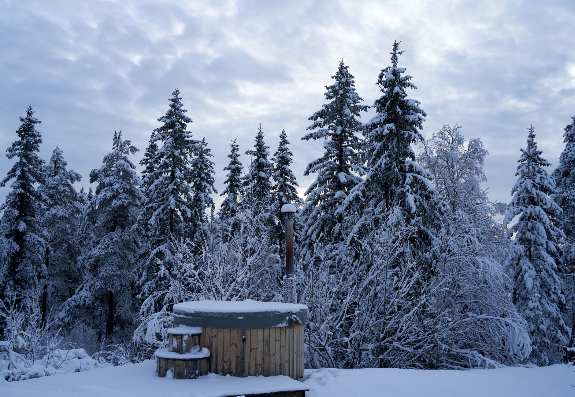 Tranquil winter forest scene with snow-covered trees and a rustic wooden hot tub, perfect for relaxation.